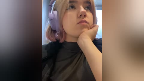 Media: Video of a young woman with fair skin, shoulder-length blonde hair, wearing purple headphones, resting her chin on her hand, wearing a black turtleneck, indoors with blurred background.