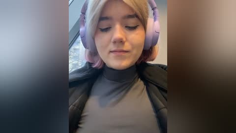 Media: Video of a young woman with fair skin, short blonde hair, wearing purple headphones and a black jacket, seated indoors with blurred background.