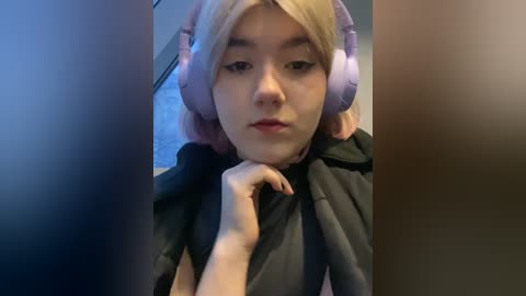 Media: Video of a young Asian woman with fair skin, blonde hair, and large pink headphones, wearing a black hoodie, looking serious and contemplative.