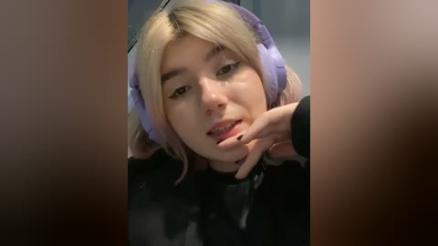 Media: Video of a young woman with light skin and blonde hair, wearing large purple headphones, lying on a dark surface with her hand near her mouth, looking introspective.