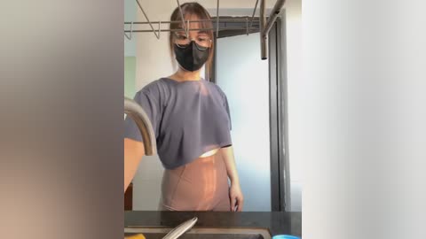 Media: Video of an East Asian woman with a slender physique, wearing a black face mask, gray T-shirt, and high-waisted, light brown pants, standing in a modern kitchen with stainless steel appliances and glass cabinets.