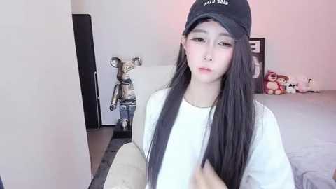 Media: Video of an Asian woman with long black hair, wearing a white t-shirt, black cap, and makeup, standing in a room with plush toys and a white bed.