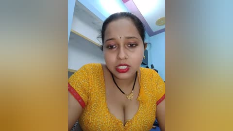 Media: A video of a South Asian woman with dark skin and dark hair, wearing a yellow polka-dotted blouse with red trim, red lipstick, and a black necklace, looking directly at the camera.