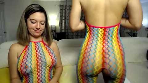 Media: Video of a fair-skinned woman with shoulder-length blonde hair wearing a vibrant, colorful, zigzag-patterned halter top that reveals her bare back and buttocks. She stands indoors near a white couch and a window with light filtering in.