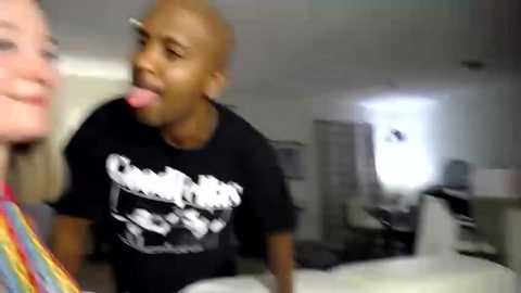 Media: Video of a young, bald Black man wearing a black T-shirt with white text, sticking out his tongue in a playful manner. He is indoors, with blurred backgrounds of furniture and a desk.