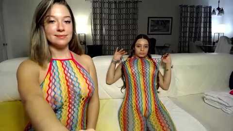 Media: Video of a young woman with fair skin and long, wavy blonde hair, wearing a colorful, zigzag-patterned halter dress, standing in a modern living room with white couch, black curtains, and framed artwork.