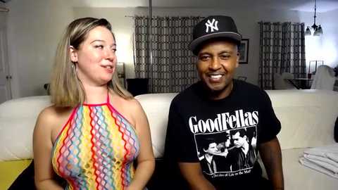 Media: Video of a light-skinned woman with blonde hair in a colorful, patterned halter top and a dark-skinned man in a black \"Goodfellas\" t-shirt and cap, sitting on a white couch in a modern, minimalist living room.