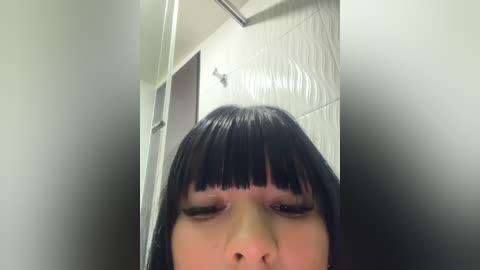 Media: A video of a young woman with straight black hair and blunt bangs, eyes closed, taken from a low angle in a bathroom with white, textured tiles.