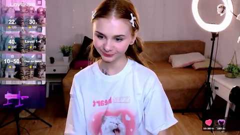 Media: Video of a young woman with fair skin and brown hair, wearing a white t-shirt featuring a pink cat graphic, smiling in a cozy, dimly lit room with a large, round light.