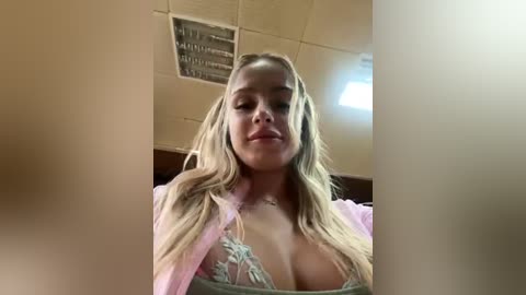 Media: Video of a blonde woman with light skin, wearing a sheer pink robe, revealing a lace bra with large breasts. The background features a beige ceiling with fluorescent lights.