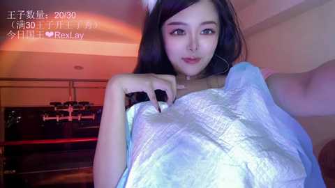 Media: Video of an East Asian woman with long dark hair, wearing a light blue sequined dress, posing seductively indoors, holding a phone camera.