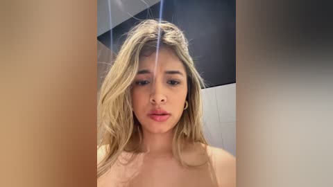 Media: Video of a young, light-skinned woman with long, wavy blonde hair, wearing a beige top, standing indoors. She has a neutral expression. Background features a modern, minimalist room with dark and light grey tones.