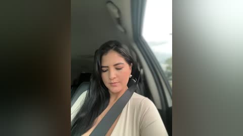 Media: A video of a woman in a car, with long black hair, wearing a beige cardigan and a seatbelt, sitting with a neutral expression.