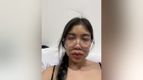 Media: Video of an Asian woman with long black hair in braids, wearing clear glasses and a black tank top, sitting on a white bed, against a plain white wall.