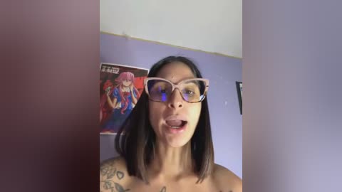 Media: Video of a young woman with medium-dark skin, straight black hair, wearing glasses, and a tattoo on her left shoulder, standing in a room with a poster of a superhero in the background.