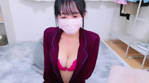 Media: Video of an Asian woman with black hair, wearing a pink face mask, a purple jacket, and a revealing pink bra, kneeling on a grey mat in a minimalist room with white shelves and clothing.