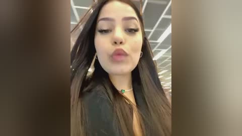 Media: Video of a young Latina woman with long, straight dark hair and a light brown complexion. She wears a black jacket and a green necklace. Background shows a modern office with a tiled ceiling and fluorescent lighting.