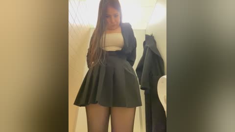 Media: Video of a young woman with long brown hair in a black pleated skirt, white top, and jacket, standing in a dimly lit bathroom.