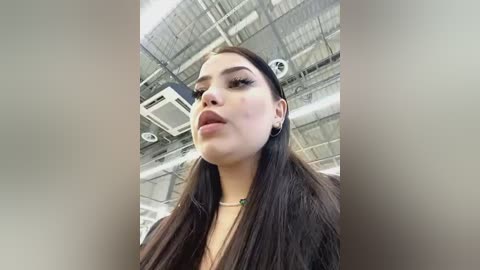 Media: Video of a young woman with long dark hair, fair skin, and full lips, wearing a black top, captured from a low angle in a warehouse with exposed ceilings and ductwork.