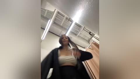 Media: A video of a young woman with long brown hair, wearing a black cardigan over a white tank top and high-waisted black pants, captured in a dimly lit, minimalist room with white walls and industrial-style ceiling lights.