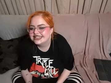 Media: Video of a smiling, fair-skinned woman with bright orange hair and glasses, wearing a black \"Trick or Treat\" T-shirt and black and white striped pants, seated on a beige couch.