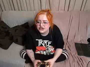 Media: Video of a plus-sized, fair-skinned woman with red hair, wearing glasses and a black \"True Blood\" t-shirt, playing a video game on a beige couch with dark pillows.