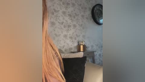 Media: A video of a woman with long, wavy blonde hair, partially visible, sitting on a black wicker chair in a room with floral-patterned wallpaper, a white wall clock, and a metallic vase on a wooden table.