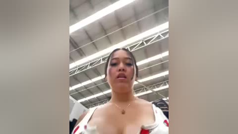 Media: Video of a young Asian woman with light skin, dark hair in a ponytail, wearing a low-cut white dress with red accents, standing in a brightly lit industrial warehouse with a high ceiling.