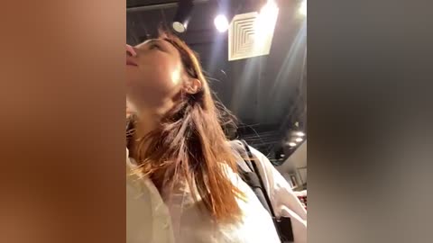 Media: Video of an Asian woman with long brown hair, wearing a white shirt, leaning back, looking upward, in a dimly lit, industrial room with exposed pipes and a ceiling fan.