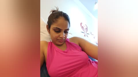 Media: A video of a South Asian woman with medium brown skin, medium build, and dark hair tied back, wearing a pink sleeveless top, lying on a bed with white sheets, in a brightly lit room with a pink heart and love quote wall art.