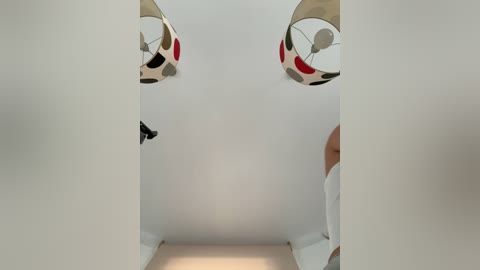 Media: Video of a minimalist, white-walled room with two spherical, abstract light fixtures featuring red and black patterns hanging from the ceiling. A person in a white shirt and black pants stands at the end of the corridor.