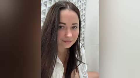 Media: Video of a young, light-skinned woman with long, straight black hair, wearing a white blouse, standing in a modern bathroom with white and geometric-patterned tiles.