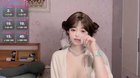 Media: Video of a young Asian woman with fair skin, dark hair styled in Princess Leia buns, wearing a white robe, standing in a cozy room with a wooden dresser, framed picture, and a green couch.