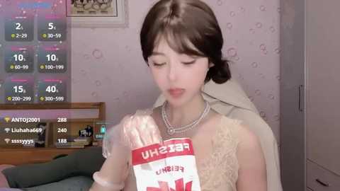 Media: A video shows a young Asian woman with fair skin and dark hair, wearing a beige lace dress, holding a pink \"Burger King\" cup. The background features a bedroom with a TV displaying a video game, and a framed photo on the wall.