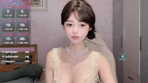 Media: Video of a young Asian woman with fair skin, long dark hair, and a slender physique, wearing a lace bodysuit with a plunging neckline. She's in a softly lit bedroom with pastel wallpaper and a framed picture on the wall.