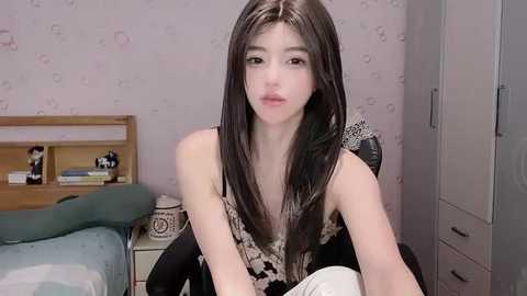 Media: A video of a young woman with long dark hair and fair skin, wearing a black and white floral sleeveless top, sitting on a bed with a white and blue comforter in a pastel-painted room with a white dresser and nightstand.