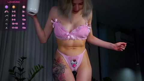 Media: Video of a slender, fair-skinned woman with blonde hair, wearing a pink lingerie set with a bow accent, holding a toilet paper roll. She has a tattoo on her thigh. The background features dimly lit room with curtains and potted plants.