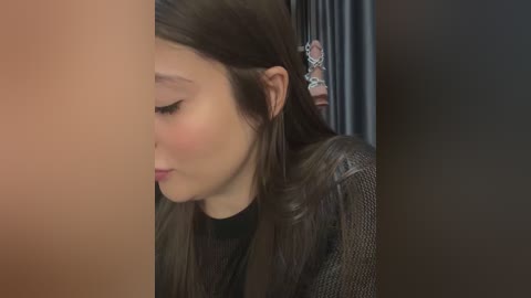 Media: Video of a young woman with long, straight brown hair, wearing a black top and a grey textured jacket, looking downward in a dimly lit room with a doll on a shelf in the background.