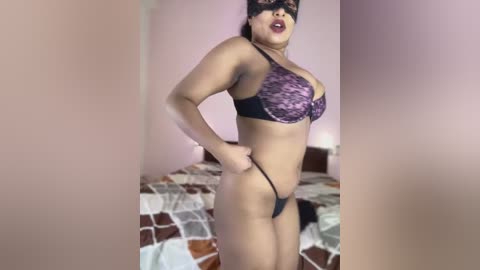 Media: Video of a curvy Latina woman in black lace lingerie, wearing a black mask, posing confidently with one hand on her hip, in a bedroom with a geometric-patterned bedspread.