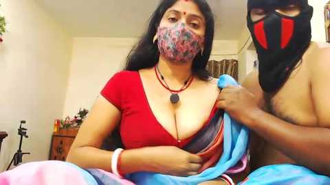 Media: Video of a topless South Asian woman with medium skin tone, large breasts, and a red sari, wearing a face mask. A man with a black mask and a blue cloth partially covers her.