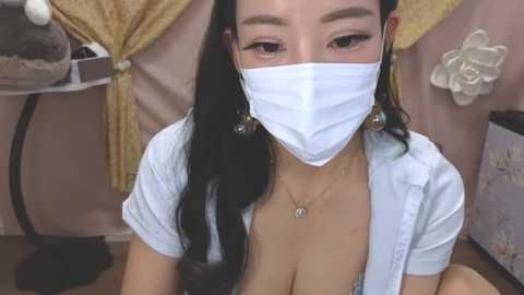 Media: Video of an East Asian woman with long black hair, wearing a white face mask, open white shirt revealing cleavage, and a delicate necklace. Background includes beige curtains and floral decor.