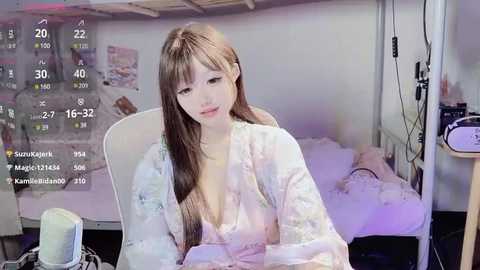 Media: A video of a young woman with long, light brown hair, wearing a white robe, sitting in a chair in a dimly lit bedroom with a bed and nightstands visible in the background.