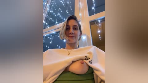 Media: A video of a young woman with short blonde hair, partially lifting her white sweatshirt to reveal her green top and left breast. She has fair skin and a relaxed expression. Background shows a cozy room with string lights and large windows.