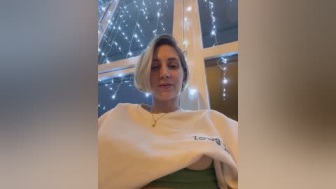 Media: A video of a young woman with short blonde hair, wearing a white sweatshirt and green top, indoors with string lights in the background.