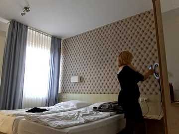 Media: A video of a woman with blonde hair, wearing a black dress, adjusting the blinds in a modern, beige-and-white bedroom with a diamond-patterned wallpaper.