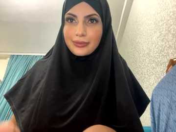 Media: Video of a young woman with olive skin, wearing a black hijab, smiling slightly. Background shows a teal curtain and textured white wall with floral pattern.