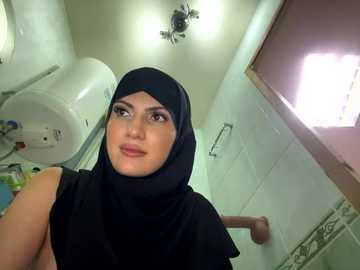 Media: Video of a young woman in a bathroom, wearing a black hijab and sleeveless top, standing next to a white toilet and tiled walls.