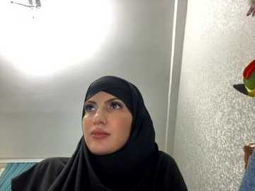 Media: Video of a woman in a black hijab, standing in a brightly lit room with a textured white wall and colorful decorations on the right.