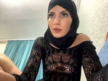 Media: Video of a woman with fair skin and dark hair in a black hijab, wearing a sheer black lace top, looking serious in a modern bedroom with light walls and blue curtains.