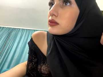 Media: Video of a Middle Eastern woman with fair skin and blue eyes, wearing a black lace top and matching hijab, against a teal curtain background.